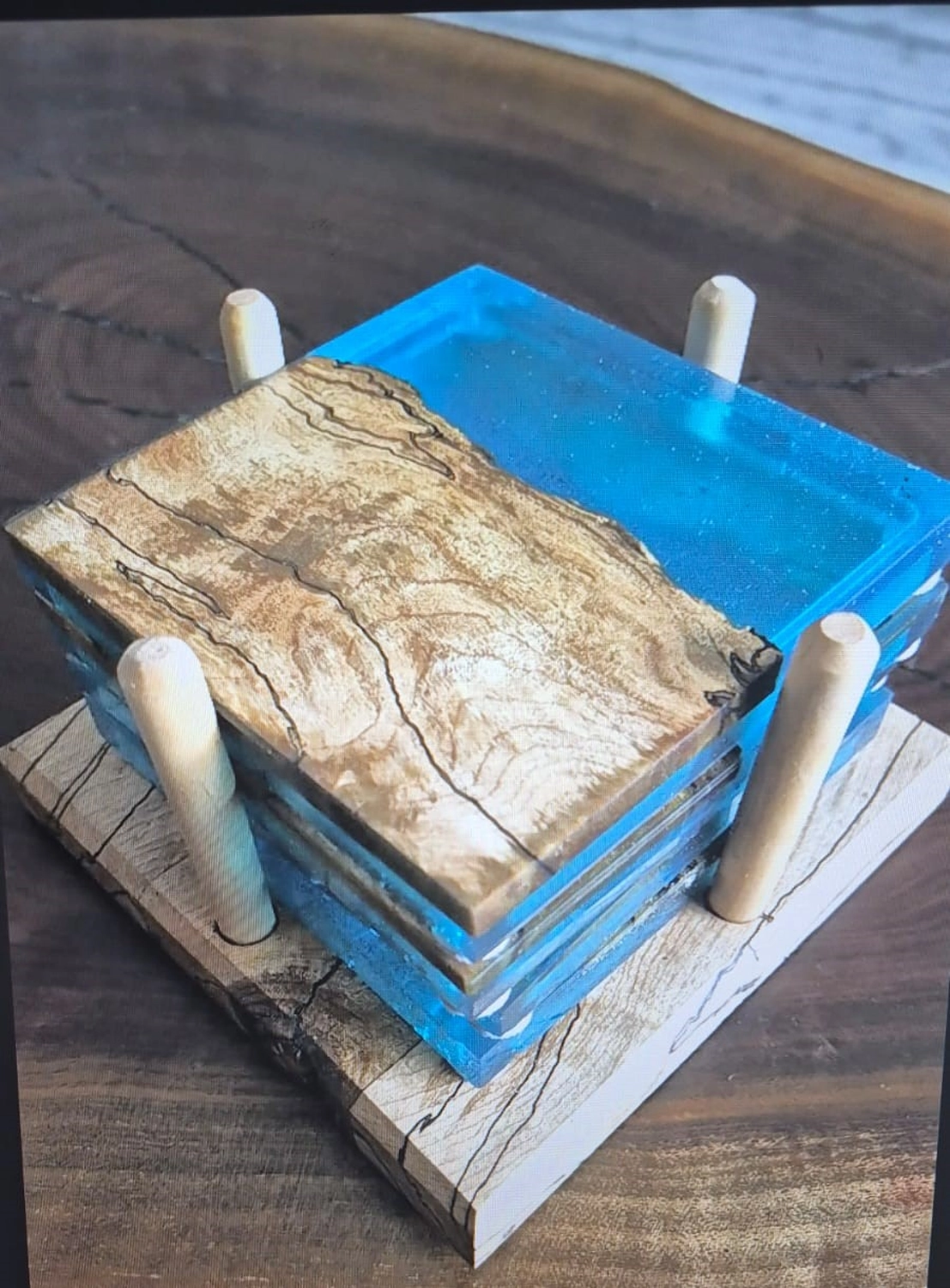Dach,Wand und Holzkonstruktion - Handgefertigte Holzkunst - Ein Set quadratischer Untersetzer aus naturbelassenem Holz und leuchtend blauem Epoxidharz, aufbewahrt in einem Holzhalter.