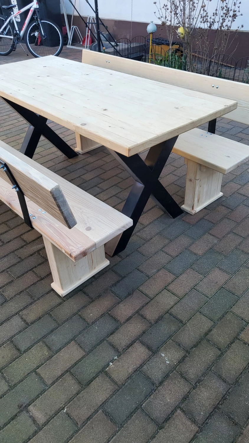 Dach,Wand und Holzkonstruktion - Handgefertigte Holzkunst - Eine rustikale Picknicktischgarnitur aus hellen Holzplanken mit dunklen, X-förmigen Metallbeinen auf einer Ziegelterrasse.