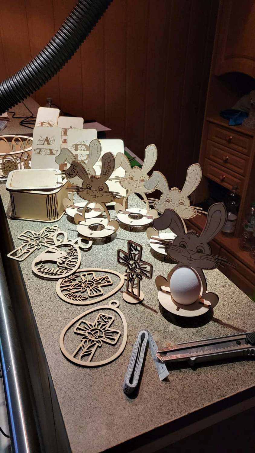 Dach,Wand und Holzkonstruktion - Handgefertigte Holzkunst - Eine Sammlung kleiner, lasergeschnittener Holzobjekte, darunter Hasenfiguren und filigrane Dekorationen, auf einer Werkbank.