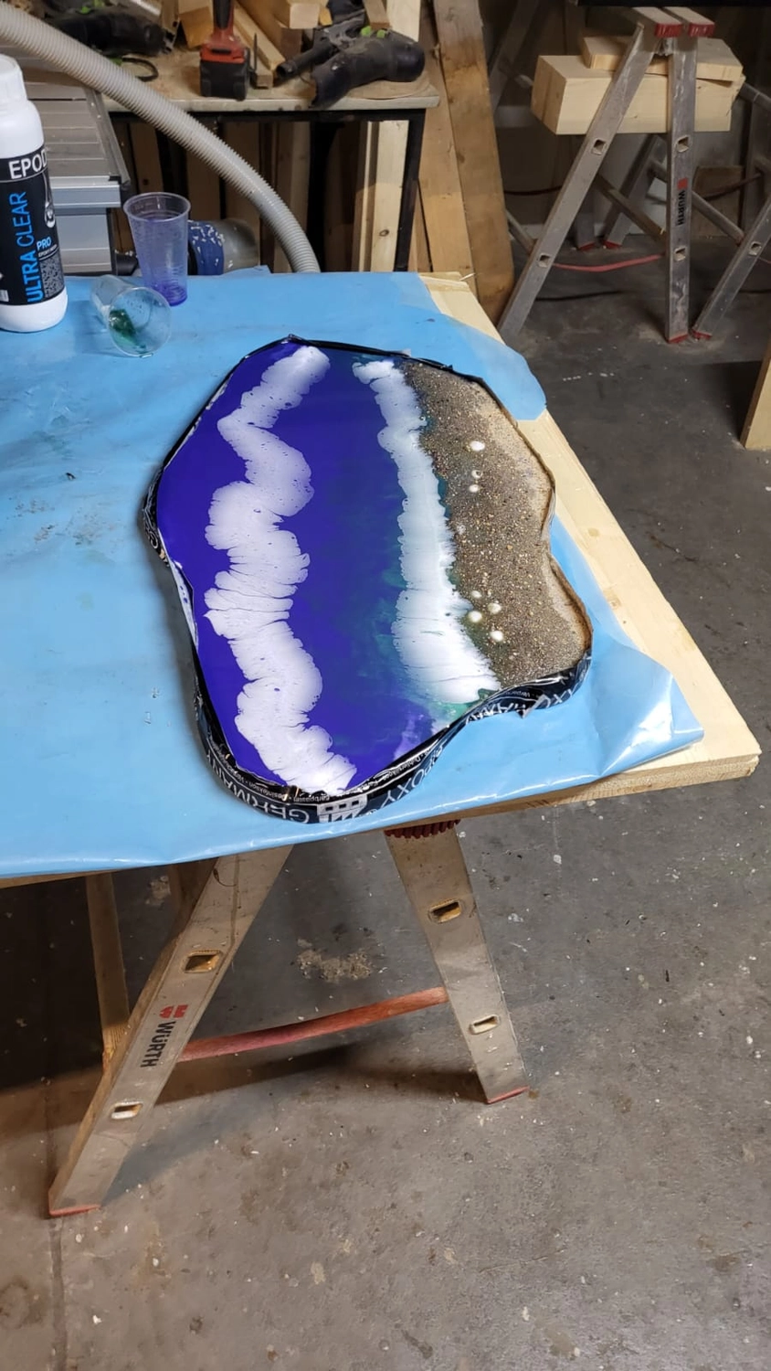 Dach,Wand und Holzkonstruktion - Handgefertigte Holzkunst - Ein lila-weißes Kunstobjekt aus Kunstharz mit geodenartigem Design auf einem blauen Werkstatttisch.