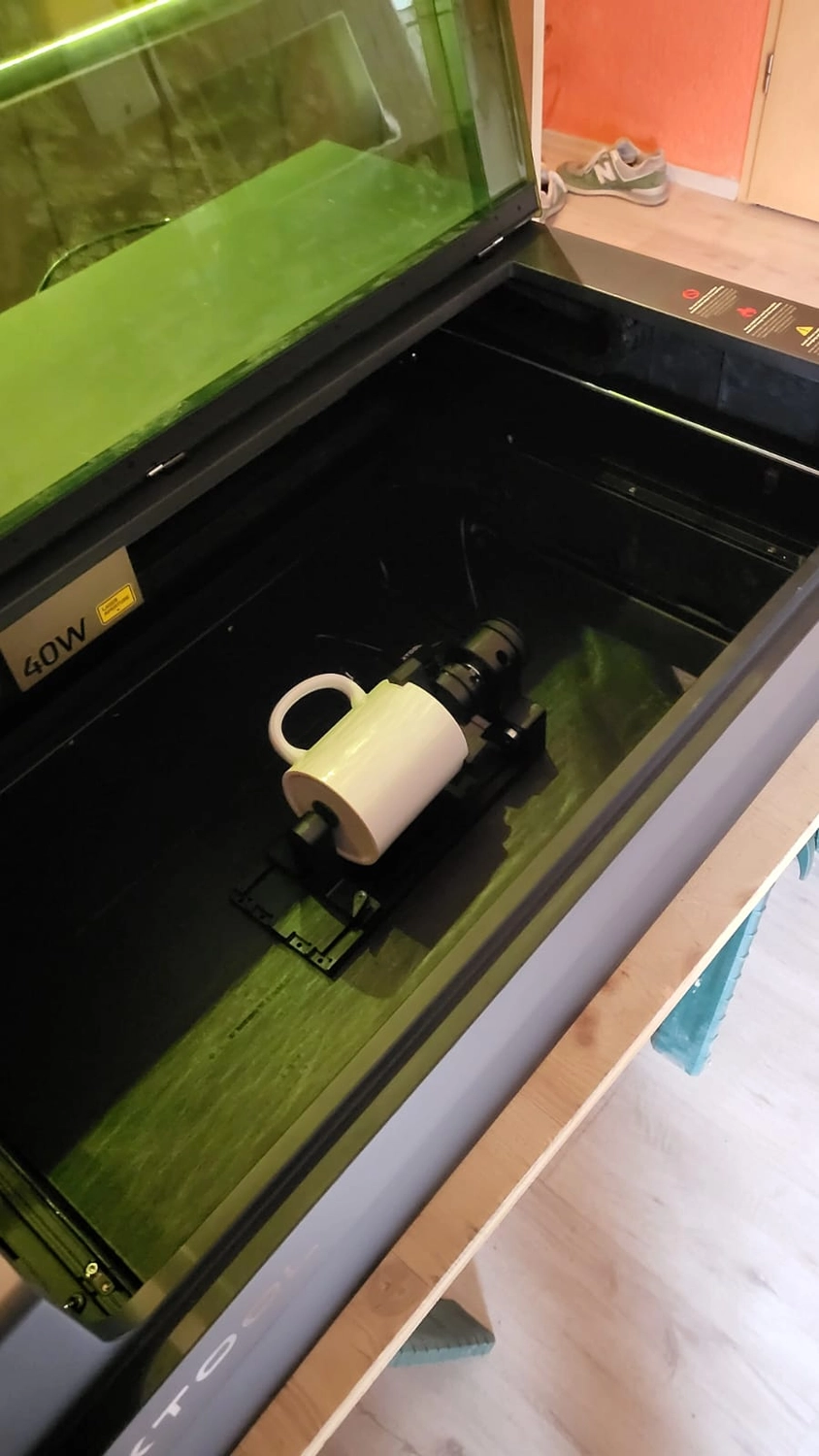 Dach,Wand und Holzkonstruktion - Handgefertigte Holzkunst - Eine Lasergravurmaschine mit 40-Watt-Etikett bearbeitet eine weiße Keramiktasse mit einem Rotationsaufsatz.