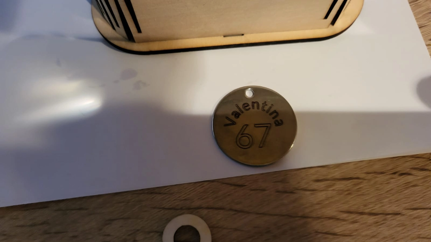 Dach,Wand und Holzkonstruktion - Handgefertigte Holzkunst - Nahaufnahme eines kleinen, runden Metallschildes mit der Gravur „Valentina“ und der Zahl „67“, das auf einer weißen Oberfläche liegt.
